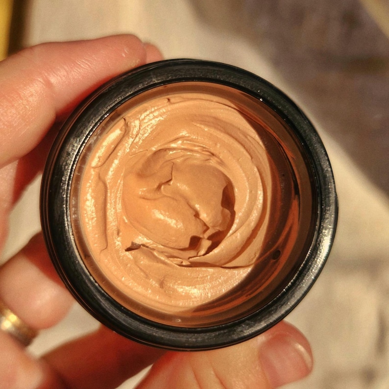 May include: A small, round, black jar filled with a thick, light brown, creamy substance. The cream has a swirled, textured surface with visible peaks and valleys, contained within a dark, circular rim that frames the product clearly.