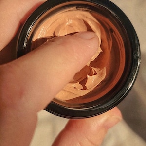 May include: A small, round, black cosmetic jar containing a thick, light brown, creamy substance with a smooth, dense texture. The product has a visible indentation from a finger pressed into the center of the cream, revealing its soft consistency.