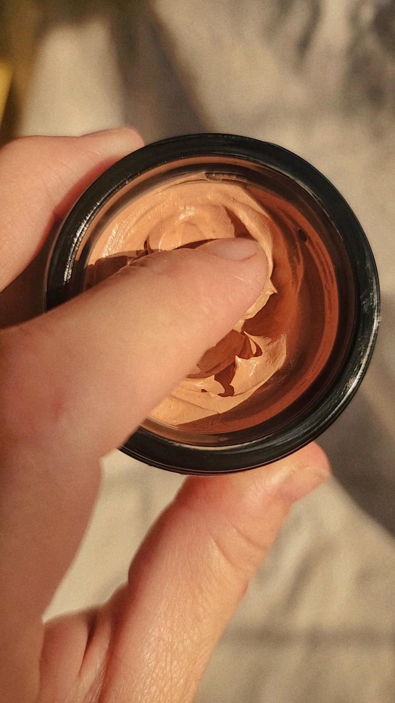 May include: A small, round, black cosmetic jar containing a thick, light brown, creamy substance with a smooth, dense texture. The product has a visible indentation from a finger pressed into the center of the cream, revealing its soft consistency.
