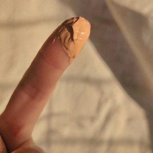 May include: A thick, light tan liquid cosmetic product with a smooth, creamy texture applied to the tip of a finger. The substance has a matte finish and appears opaque, showing a consistent beige color throughout the dollop of product.