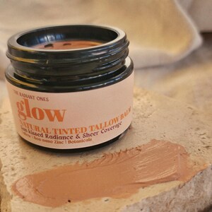 May include: A small black jar of The Radiant Ones glow natural tinted tallow balm with a beige label featuring black text. A smear of the tan-colored balm is visible on a textured stone surface in front of the open jar, showing the product texture.