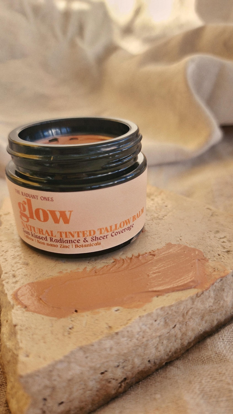 May include: A small black jar of The Radiant Ones glow natural tinted tallow balm with a beige label featuring black text. A smear of the tan-colored balm is visible on a textured stone surface in front of the open jar, showing the product texture.