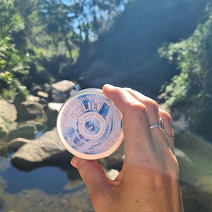 May include: A round, silver tin of "RELIEF" cream is held up in front of a blurred natural background. The tin's label includes text such as "Oatmeal Manuka" and "Chamomile". The cream is marketed as "The Radiant Ones".