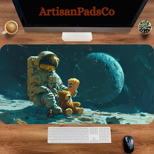 Op de afbeelding: Een rechthoekige muismat met een astronaut op een maan, die popcorn en een teddybeer vasthoudt. De achtergrond toont een planeet en sterren. De mat heeft bovenaan de tekst "ArtisanPadsCo".