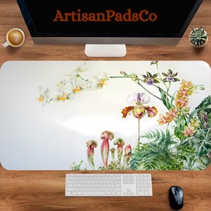May include: A white desk mat featuring a vibrant floral design with orchids and other exotic flowers. The mat has the text "ArtisanPadsCo" at the top. A keyboard, mouse, and coffee cup are visible on a wooden desk.
