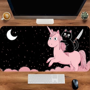 May include: A black desk mat featuring a cartoon unicorn with a pink mane and a black cat holding a knife. The background is black with a crescent moon, stars, and pink clouds. A white keyboard and mouse are visible.