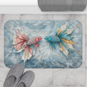 May include: A rectangular bath mat featuring two stylized betta fish facing each other. One fish is pink and white, the other is blue and yellow, set against a blue and white watercolor background. Gray slippers are in the foreground.
