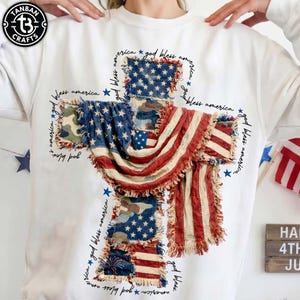 May include: White sweatshirt featuring a cross design made from the American flag. The cross is composed of patches of the flag, with frayed edges. The words "God Bless America" and stars are printed around the cross. The brand logo is in the upper left corner.