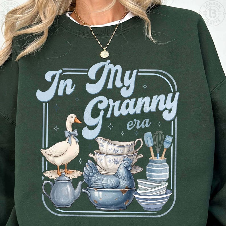 May include: Dark green sweatshirt with the text "In My Granny era" in light blue. The graphic includes a goose with a blue bow, teacups, a chicken, and kitchen utensils. The design has a vintage, farmhouse aesthetic.