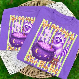 May include: A purple t-shirt featuring a graphic of a raccoon stirring a purple cauldron with a wooden spoon. The design includes light green vertical stripes, stars, a crescent moon, and the text On my way to Stir The Pot in orange.