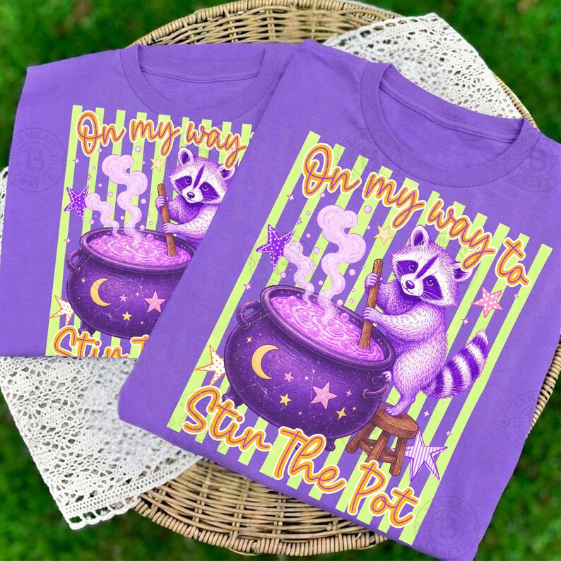 May include: A purple t-shirt featuring a graphic of a raccoon stirring a purple cauldron with a wooden spoon. The design includes light green vertical stripes, stars, a crescent moon, and the text On my way to Stir The Pot in orange.