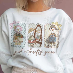Puede incluir: Sudadera blanca con tres ilustraciones enmarcadas de un ganso con diferentes atuendos. El ganso se representa con un sombrero floral, un espejo y un carrito de la compra. El texto "just a thrifty goose" est&aacute; escrito debajo.