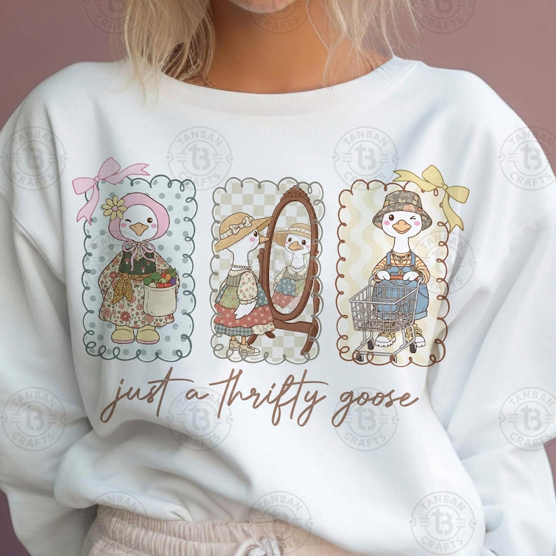 Puede incluir: Sudadera blanca con tres ilustraciones enmarcadas de un ganso con diferentes atuendos. El ganso se representa con un sombrero floral, un espejo y un carrito de la compra. El texto "just a thrifty goose" est&aacute; escrito debajo.