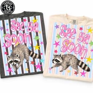 May include: Two t-shirts, one dark gray and one cream, each featuring a raccoon graphic. The design includes the text "See ya Soon" in pink bubble letters, set against a striped background with colorful stars.