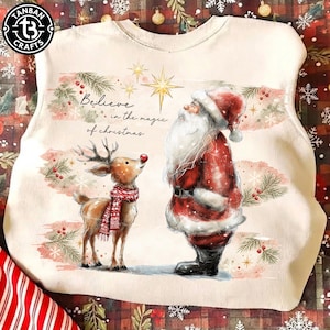 May include: A cream-coloured sweatshirt featuring a graphic of Santa Claus and a reindeer wearing a red scarf. The text reads, "Believe in the magic of Christmas." The design includes a gold star, pine branches, and red berries on a light background.