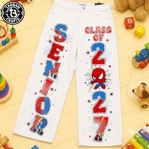 May include: White denim jeans featuring a vertical graphic print on each leg. The left leg displays the word SENIOR in red and blue block letters. The right leg features the text CLASS OF 2027 with a cartoon spider character and spiderwebs.