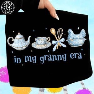 May include: Black tote bag with a blue and white polka-dot design featuring a teapot, teacup, whisk, spoon, and a chicken in a bowl. The words "in my granny era" are printed in blue. The bag is held by a person.