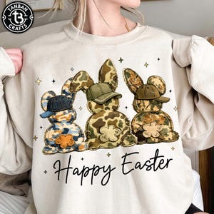Op de afbeelding: Beige sweatshirt met drie konijnenfiguren met camouflagepatronen en baseballpetten. De konijnen hebben bloemaccenten en zijn in een rij gerangschikt. De woorden "Happy Easter" staan eronder in een script.