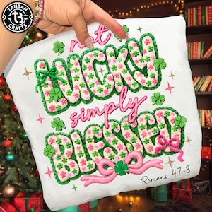 Puede incluir: Cojín blanco con el texto bordado "Not Lucky Simply Blessed" en verde y rosa, adornado con tréboles y lazos. También incluye el texto "Romanos 4:7-8". Un artículo temático del Día de San Patricio.