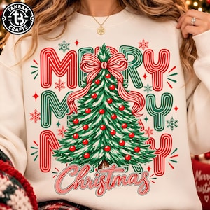 May include: A cream-colored sweatshirt featuring a central graphic of a green Christmas tree adorned with red ornaments and a large red and white striped bow. The text MERRY CHRISTMAS is printed in a stylized red font around the tree.