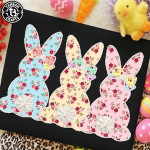 May include: Three Easter bunny decorations in light blue, yellow, and pink, each adorned with a floral pattern and a decorative bow. The bunnies have pearl-like trim and a white, circular tail. The background is black.