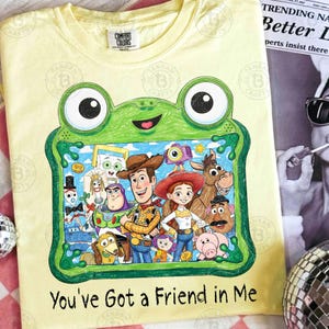 May include: A pale yellow t-shirt featuring a green frog-shaped frame graphic containing a colorful illustration of various animated characters. Below the frame, the text reads, You've Got a Friend in Me. The shirt has a Comfort Colors brand label.