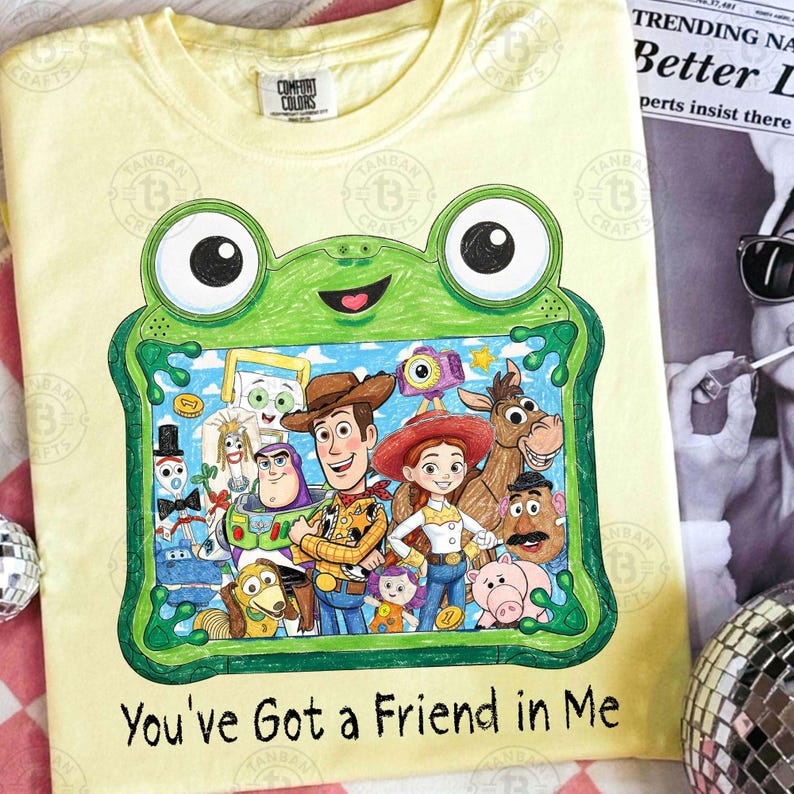 May include: A pale yellow t-shirt featuring a green frog-shaped frame graphic containing a colorful illustration of various animated characters. Below the frame, the text reads, You've Got a Friend in Me. The shirt has a Comfort Colors brand label.