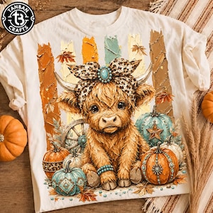 May include: A cream-colored t-shirt featuring a graphic of a fluffy Highland cow wearing a leopard print bow with a turquoise stone. The cow is surrounded by pumpkins, a wagon wheel, and autumn leaves against vertical textured paint strokes.