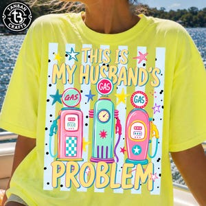 Pode incluir: Camiseta amarela com o texto "THIS IS MY HUSBAND'S PROBLEM" e ilustrações de bombas de gasolina vintage em rosa, verde-azulado e verde. O design inclui estrelas e listras.