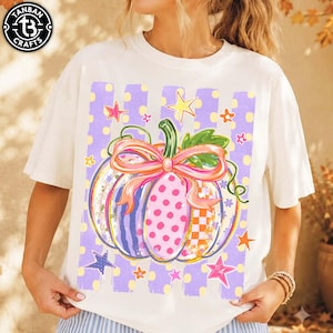 May include: A white t-shirt featuring a central graphic of a pumpkin with multicolored segments, including polka dots, stripes, and checkered patterns. The pumpkin is adorned with a large pink bow and green leaves, set against a light purple striped background with yellow dots and various colorful stars.