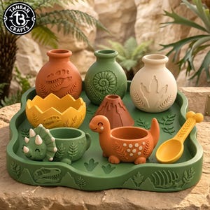 May include: A set of dinosaur-themed silicone kitchenware including three small vases, a yellow cracked egg bowl, a green triceratops bowl, an orange dinosaur bowl, a brown volcano, and a yellow spoon, all arranged on a green tray with fossil motifs.