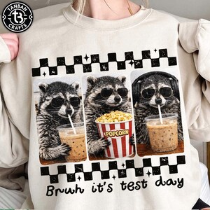 May include: Cream-coloured sweatshirt with a graphic of three raccoons wearing sunglasses. One raccoon sips a drink, another holds popcorn, and the third wears headphones. The text "Bruh it's test day" is below the graphic.