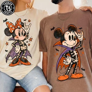 Może przedstawiać: Dwie koszulki z grafikami o tematyce Halloween. Kremowa koszulka przedstawia postać Myszki Minnie w kostiumie szkieletu z peleryną i pomarańczową kokardą. Brązowa koszulka przedstawia Myszkę Miki w kostiumie szkieletu z fioletową peleryną.