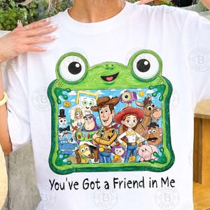 May include: A white t-shirt featuring a green frog-shaped frame graphic containing a colorful illustration of various Toy Story characters. Below the graphic, the text reads, "You've Got a Friend in Me" in a black, handwritten-style font.