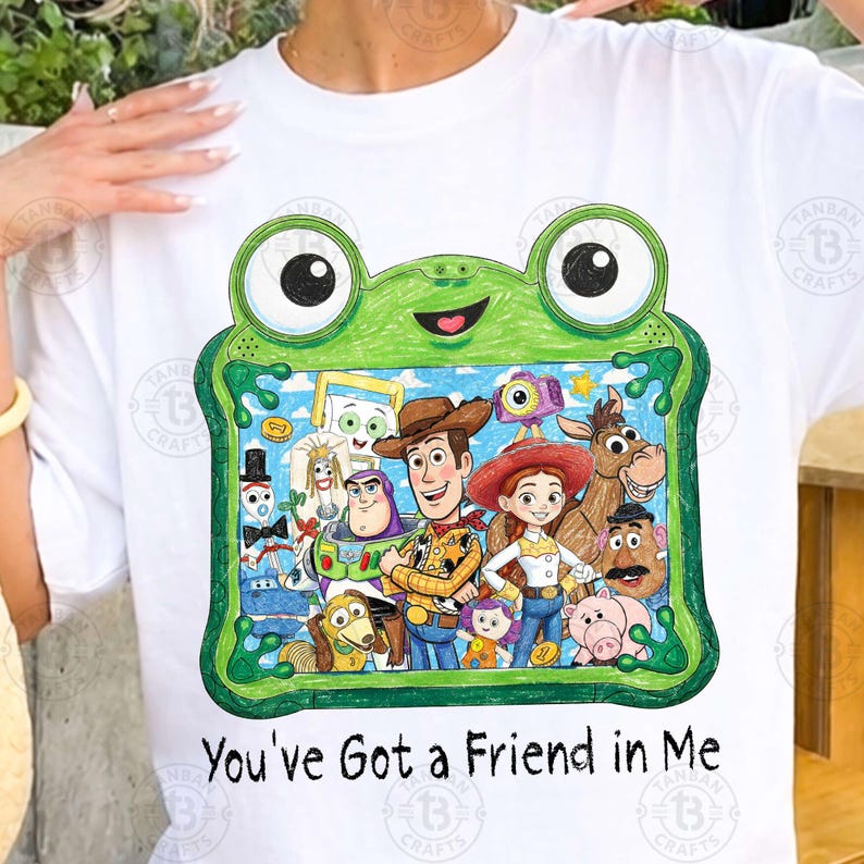 May include: A white t-shirt featuring a green frog-shaped frame graphic containing a colorful illustration of various Toy Story characters. Below the graphic, the text reads, "You've Got a Friend in Me" in a black, handwritten-style font.