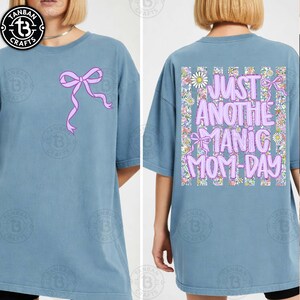 May include: A light blue t-shirt with a pink bow graphic on the front. The back of the shirt features the text "JUST ANOTHER MANIC MOM-DAY" in pink, surrounded by flowers and bows. The shirt is a casual, oversized style.