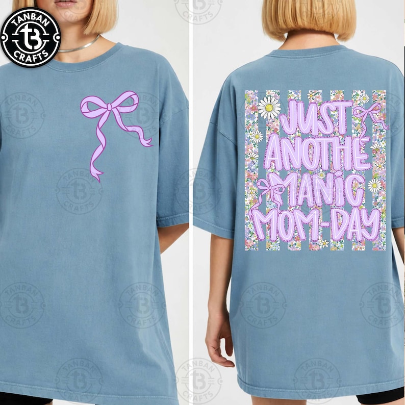 May include: A light blue t-shirt with a pink bow graphic on the front. The back of the shirt features the text "JUST ANOTHER MANIC MOM-DAY" in pink, surrounded by flowers and bows. The shirt is a casual, oversized style.