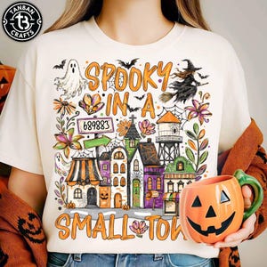 May include: A cream-colored t-shirt featuring a colorful Halloween-themed graphic with the text "SPOOKY IN A SMALL TOWN" and the number "689883". The design includes a ghost, a witch on a broom, a water tower, and various small buildings.