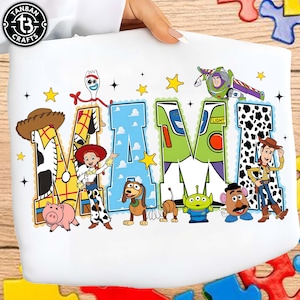 Puede incluir: Camiseta blanca con la palabra "MAMA" en un diseño colorido con personajes de Toy Story. Las letras están decoradas con imágenes de Woody, Buzz Lightyear y otros personajes. La camiseta está sujeta por una persona.