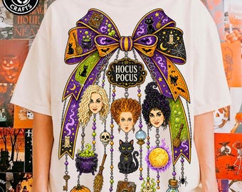 May include: A cream-colored t-shirt featuring a large graphic of a purple and orange bow with the text Hocus Pocus. The design includes three women's faces, a black cat, a pumpkin, a cauldron, a spellbook, a broom, and various magical icons.