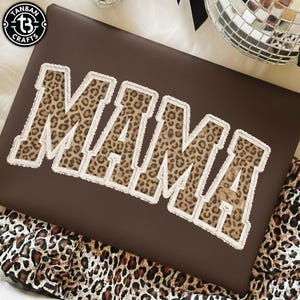 May include: A brown pillow with the word "MAMA" in large, leopard print letters with a white, fringed border. The pillow is decorated with a leopard print pattern.