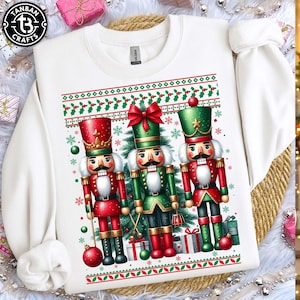May include: White sweatshirt with a festive design of three nutcracker figures in red and green. The design includes a Christmas tree, snowflakes, and decorative borders with holly and berries. The sweatshirt has long sleeves.