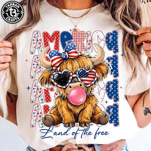 Puede incluir: Camiseta color crema con una vaca de las Tierras Altas de dibujos animados con gafas de sol en forma de corazón y una burbuja de chicle. La palabra "AMERICA" está impresa en una fuente patriótica, y la frase "Land of the free" está escrita debajo.