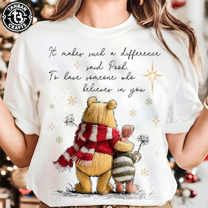 May include: White t-shirt with a graphic of Winnie the Pooh and Piglet embracing, Pooh wearing a red and white scarf. The text says "It makes such a difference said Pooh. To have someone who believes in you."