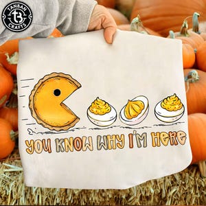 Può includere: Un canovaccio bianco con un design umoristico. Raffigura una fetta di torta come Pac-Man, seguita da uova alla diavola. Il testo "YOU KNOW WHY I'M HERE" è stampato sotto. Lo sfondo mostra zucche.