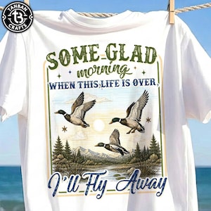 以下が含まれることがあります： 自然をテーマにしたグラフィックが施された白いTシャツ。湖、木、山のある風景の上を飛ぶ3羽のアヒルがデザインされています。「SOME GLAD morning WHEN THIS LIFE IS OVER, I'll Fly Away」というテキストが含まれています。