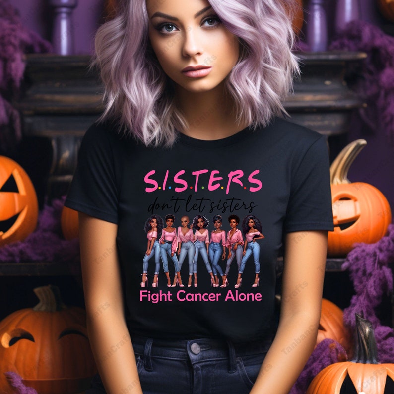 May include: Black t-shirt with the text "S.I.S.T.E.R.S don't let sisters Fight Cancer Alone". The shirt features a graphic of six women in pink tops and blue jeans. Pumpkins and purple decor are in the background.