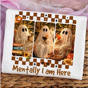 May include: White t-shirt with a whimsical design featuring three ghost figures enjoying leisure activities. The ghosts are depicted with black oval eyes and draped in white sheets. The text "Mentally I am Here" is printed in brown.