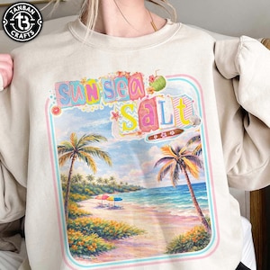 May include: Beige sweatshirt featuring a colorful beach scene with palm trees, ocean, and the words "Sun Sea Salt." The design includes a retro frame and tropical elements, evoking a vacation vibe. The sweatshirt has a crew neck.