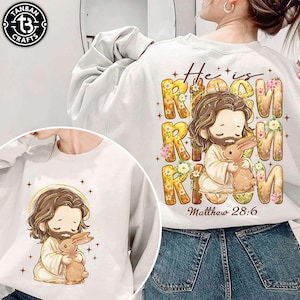 May include: White sweatshirt featuring a cartoon illustration of Jesus holding a brown bunny. The text "He is Risen" is displayed in a floral design, with "Matthew 28:6" below. The sweatshirt has a crew neck.